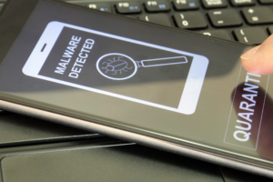 A smartphone displaying a warning message that reads "MALWARE DETECTED" alongside an icon of a magnifying glass over a bug. The phone is positioned over a laptop keyboard, and a finger is pressing the "QUARANTINE" button on the screen.