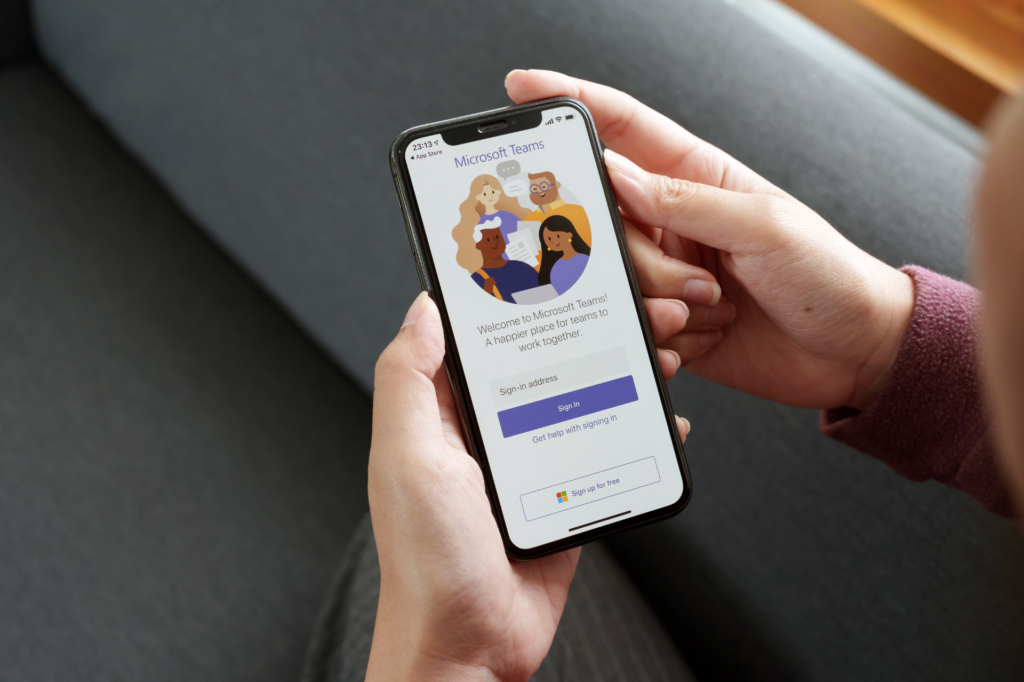 A person holding a smartphone displaying the Microsoft Teams app on the screen. The app's welcome screen reads, "Welcome to Microsoft Teams! A happier place for teams to work together," with options to sign in or sign up.