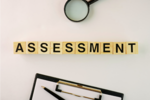 Wooden blocks spelling "ASSESSMENT" with a magnifying glass above and a clipboard and pen below on a white background.