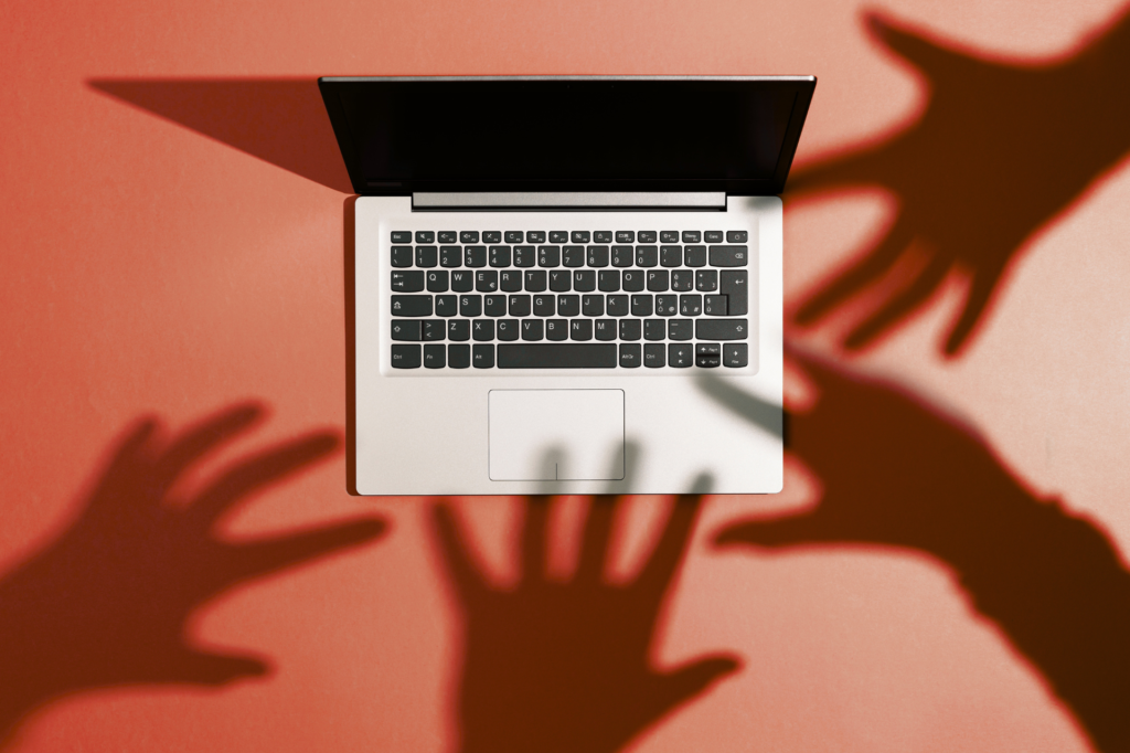 A laptop on a red background with shadows of hands reaching towards it, symbolizing potential cyber threats or data breach pitfalls to avoid.