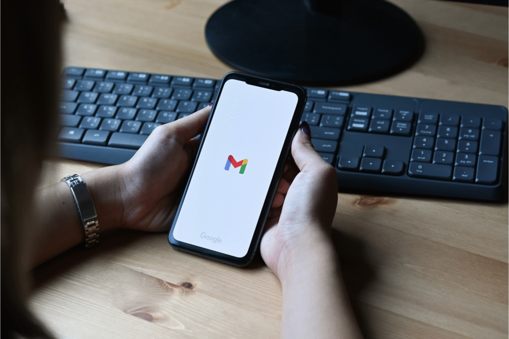 Person holding a smartphone displaying the Gmail app logo, with a keyboard and monitor in the background on a wooden desk.