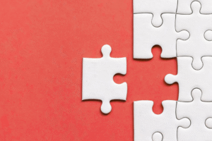 A single white puzzle piece about to be placed into a nearly completed puzzle on a red background.