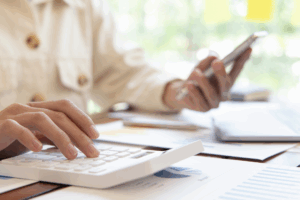 Person using a calculator and holding a smartphone while reviewing financial documents with graphs and charts.