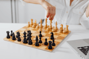 A person in a white shirt moving a chess piece on a wooden chessboard next to a laptop.