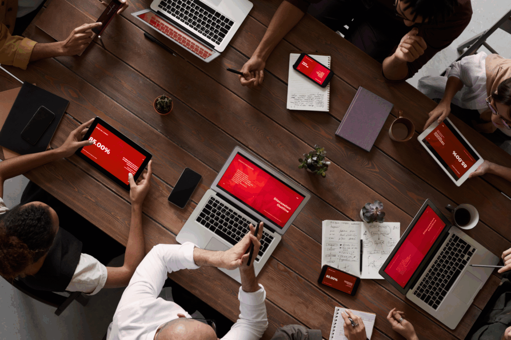 A group of people sitting around a wooden table working on laptops, tablets, and smartphones displaying red presentation slides, with notebooks, coffee cups, and small plants also on the table.