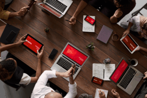 A group of people sitting around a wooden table working on laptops, tablets, and smartphones displaying red presentation slides, with notebooks, coffee cups, and small plants also on the table.